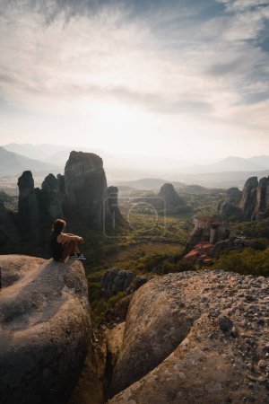 Foto de Hombre sentado disfrutando al atardecer del hermoso paisaje de los monasterios sobre rocas de Meteora. Durante su viaje turístico por Grecia - Imagen libre de derechos