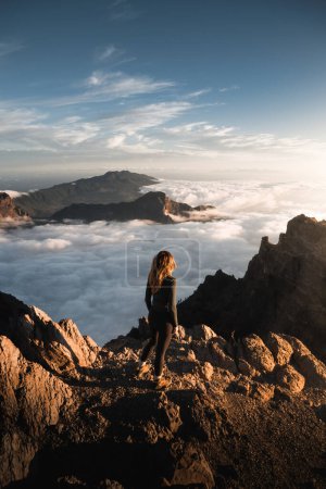 Foto de Joven mujer disfrutando de una hermosa puesta de sol desde lo alto del Roque De Los Muchachos con un mar de nubes sobre Caldera De Taburiente en la isla canaria de La Palma. - Imagen libre de derechos