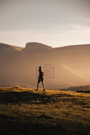 Téléchargez les photos : Jeune femme marchant au coucher du soleil à travers un champ avec le paysage du pic Ungino des montagnes Gorobel en arrière-plan - en image libre de droit