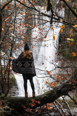 Téléchargez les photos : Jeune femme avec cape et chapeau observant une cascade en automne parmi les arbres aux feuilles rougeâtres - en image libre de droit
