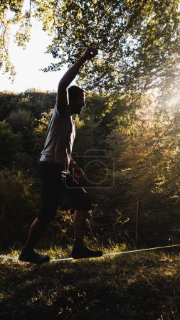 Foto de Hombre haciendo slacklining en el bosque en una puesta de sol del verano con los rayos de sol que filtran a través de las hojas de los árboles - Imagen libre de derechos