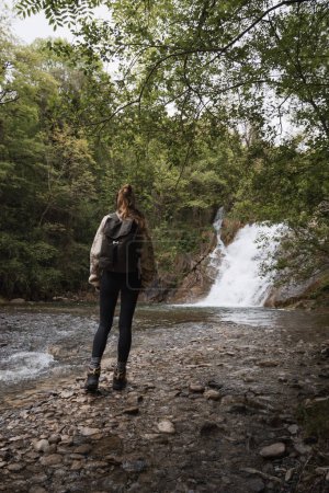 Foto de Mujer joven caminando junto a una cascada en un entorno de bosque verde - Imagen libre de derechos