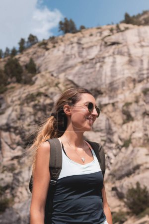Téléchargez les photos : Jeune femme aux lunettes de soleil souriant à la montagne lors de son itinéraire randonnée et aventure - en image libre de droit