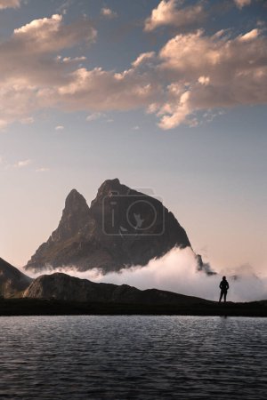 Téléchargez les photos : Paysage spectaculaire avec le majestueux pic Midi d'Ossau au-dessus des nuages au coucher du soleil et une personne profitant des vues à côté du lac Anayet. en Pyrénées - en image libre de droit