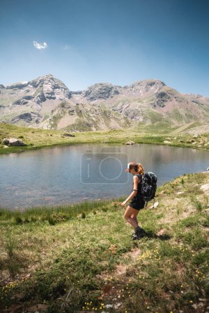 Foto de Joven mochilera trekking junto a un lago con las montañas de los Pirineos al fondo en un soleado día de verano en Formigal, Huesca. - Imagen libre de derechos