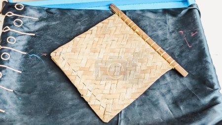 Vista de ángulo alto de un ventilador de mano tejido de bambú colocado en un escritorio de madera de color azul cubierto por una pieza de tela decorativa de color oscuro. Se utiliza comúnmente para abanicar las brasas en la estufa de carbón al cocinar. Concepto de método tradicional de cocina indonesia