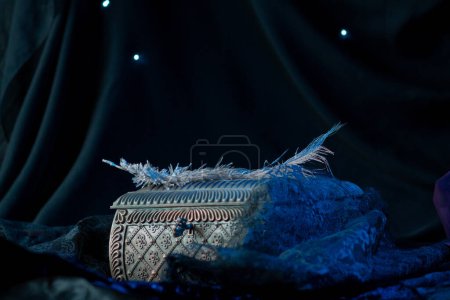 Photo for The wrought iron chest is decorated with a carved pattern, on which lie ostrich feathers in blue neon light. Magical artifact against dark drapery in a black circle - Royalty Free Image