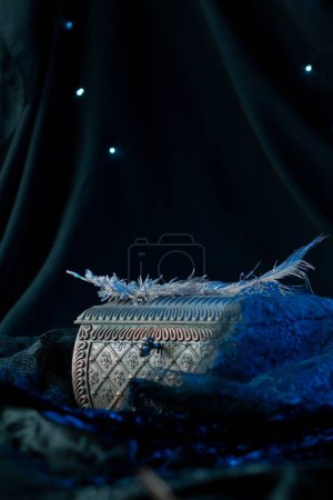 Photo for The wrought iron chest is decorated with a carved pattern, on which lie ostrich feathers in blue neon light. Magical artifact against dark drapery in a black circle, vertically - Royalty Free Image