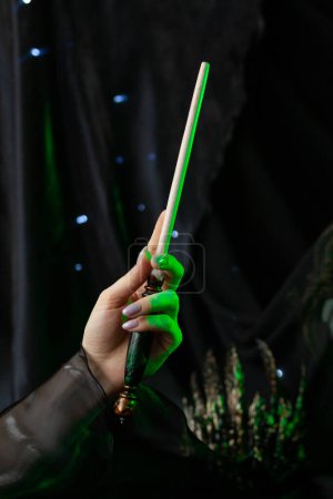 Foto de La mano de la bruja oscura, iluminada por la luz verde de neón, sostiene una varita mágica hecha de madera, decorada con un adorno sobre un fondo negro con una corona espeluznante. Artefacto mágico primer plano, vertical - Imagen libre de derechos