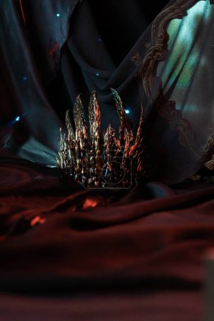 Die Schurken ominöse vergoldete Krone mit scharfen Ecken liegt auf Falten aus dunklem Tuch in der Nähe eines alten Spiegels, der mit schwarzem Chiffon bedeckt ist. Halloween-Accessoire im Rotlicht, vertikal