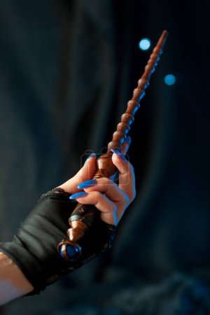 Photo for The hand of a sorceress with blue nails holds a magic wand, guiding it for a spell on a dark background with shining lights. Cosplay from Hogwarts Legacy, close-up vertically - Royalty Free Image