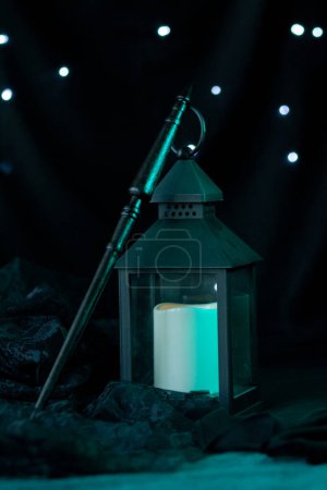 Photo for A black magic wand of a wood carver stands near an extinguished dusty lantern. Mystical magical attributes are located on a dark background with shining lights, vertically close-up - Royalty Free Image
