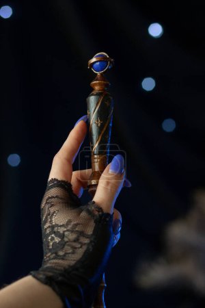 Photo for The hand of a sorceress with blue nails holds a magic wand, guiding it for a spell on a dark background with shining lights. Cosplay from Hogwarts Legacy, close-up vertically - Royalty Free Image