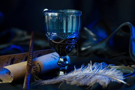 Photo for A blue glass goblet, a goblet decorated with a three-dimensional ornament, stands among magical vintage things next to Cho Changs magic wand. Halloween decor on dark fabric in neon light, close up - Royalty Free Image