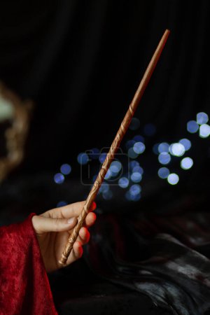 Photo for A magicians hand in a red robe holds an elegant wooden magic wand decorated with floral carvings on a dark background with blue lights. Arcane artifact close-up vertical - Royalty Free Image
