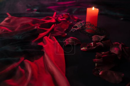 Massagekerze auf einem dunklen Bett zwischen Rosenblättern, Damenschmuck, neben einem roten Satinkleid. Vorbereitung auf ein leidenschaftliches Date am Valentinstag, Nahaufnahme