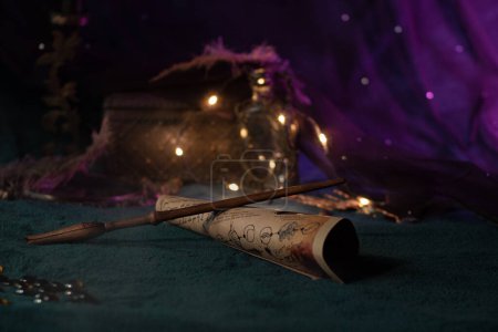 A magic wand carved from wood lies on a scroll with spells on a velvet green cloth among magical paraphernalia on a shining background with lights. Wizards Things Close-Up