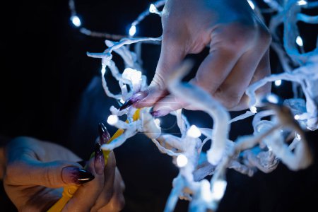 The hands of the master sculpt thin white twigs of the crown, weaving a shining garland into them on a dark background in blue neon light. The process of making a mystical accessory, close-up