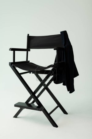 Téléchargez les photos : La chaise de réalisateur noire est pliante, sur laquelle une veste sombre est suspendue sur le côté. Fauteuil en bois sur le fond d'un cyclorama blanc dans un studio photo - en image libre de droit