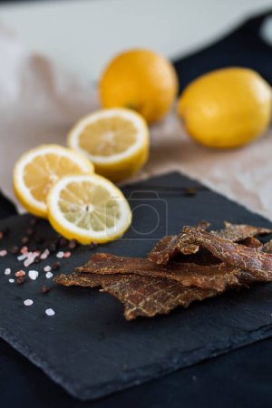 Photo for Chunks of dried turkey meat lie next to whole and sliced lemons, scattered pink salt and whole black pepper on a stone square plate. Food, snacks, spices close-up vertically - Royalty Free Image