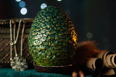 Un gran huevo verde escamoso de dragón se encuentra en un soporte junto a un cofre antiguo con un collar entre pergaminos y telas, sobre un fondo oscuro con bokeh brillante. Tesoros fabulosos, primer plano