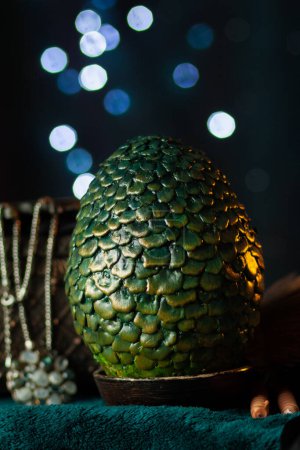 Un gran huevo verde escamoso de dragón se encuentra en un soporte junto a un cofre antiguo con un collar entre pergaminos y telas, sobre un fondo oscuro con bokeh brillante. Tesoros fabulosos, primer plano