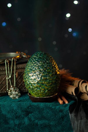 Un gran huevo verde escamoso de dragón se encuentra en un soporte junto a un cofre antiguo con un collar entre pergaminos y telas, sobre un fondo oscuro con bokeh brillante. Tesoros fabulosos, de cerca verticalmente