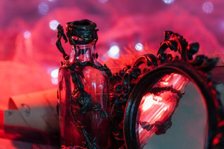 Une bouteille de forme inhabituelle, décorée d'un ornement floral sculpté, se dresse près du cadre d'un miroir vintage forgé sur un fond rouge brillant avec des guirlandes. Articles magiques pour l'intérieur