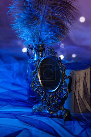 Un pequeño espejo vintage en un marco de hierro forjado se encuentra cerca de una botella de plumas de avestruz entre las luces brillantes de una guirnalda sobre un fondo azul brillante. Atmósfera mágica, primer plano vertical