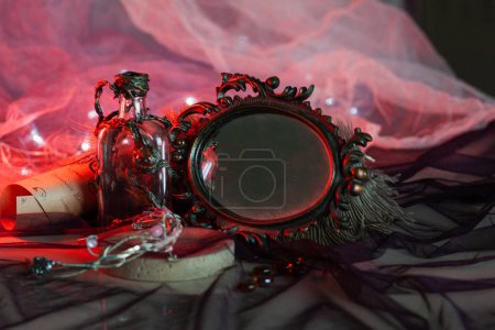 An unusually shaped bottle, decorated with a sculpted floral ornament, stands near the frame of a forged vintage mirror on a red shining background with garlands. Magical items for the interior