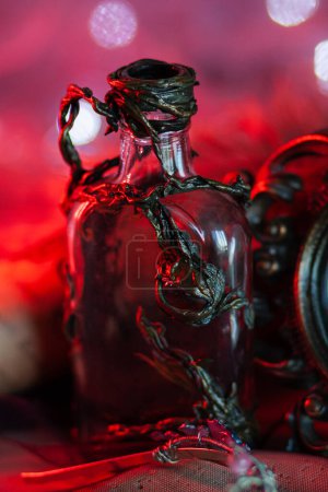 An unusually shaped bottle, decorated with a sculpted floral ornament, stands near the frame of a forged vintage mirror on a red shining background with garlands. Magical items for the interior