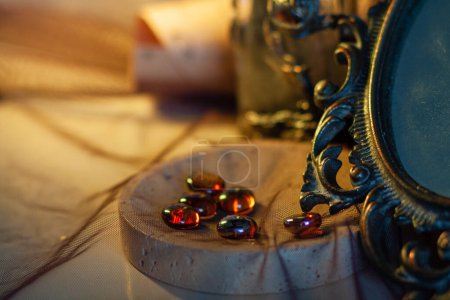 Glowing glass round stones lie in a heap in a decorative bowl next to an old vintage mirror with a curly metal frame against a warm golden light. Close-up, details