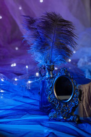 A vintage small mirror in a wrought-iron frame stands near a bottle of ostrich feathers among the shining lights of a garland on a bright blue background. Magical atmosphere, close-up vertically