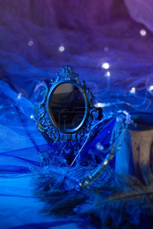 A vintage mirror in a wrought-iron frame stands next to ostrich feathers among glowing lights of a garland against a bright blue background. Magical atmosphere, vertical close-up