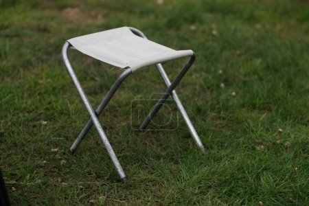 A folding camping and picnic chair with a metal frame and a light fabric seat sits on the grass. Tourist furniture vertically close-up
