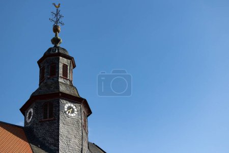 Photo for Church clock surrounded by roof shingles. Close up church tower with clock. Roff shingle tower wit ancient clock and old white, black and golden clock-face. Sunny day with blue sky and few clouds. - Royalty Free Image