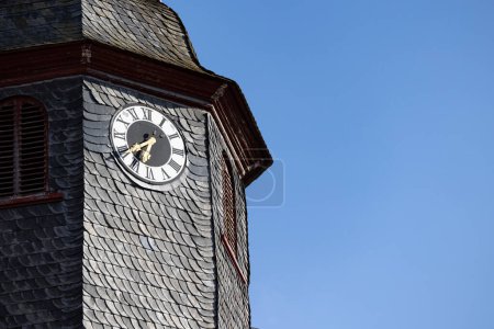 Photo for Church clock surrounded by roof shingles. Close up church tower with clock. Roff shingle tower wit ancient clock and old white, black and golden clock-face. Sunny day with blue sky and few clouds. - Royalty Free Image
