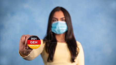 Photo for Young woman with german election button and mask. Young woman with a mask stretches out her arm and holds an election button in the German national colors, with the statement - 2021 Geh waehlen. (2021 Go Vote) - Royalty Free Image