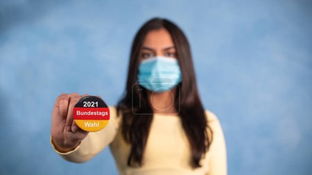 Photo for Young woman with german election button and mask. Young woman with a mask stretches out her arm and holds an election button in the German national colors, with the statement - 2021 Bundestagswahl. (2021 Federal election) - Royalty Free Image