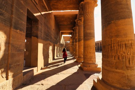 Un visitante camina por las hermosas colonadas en el patio bajo el sol dorado de la noche en el Templo de Isis en la Isla Philae en el lago Nasser, construido por Nectanebo y Ptolomeo Faroas cerca de Asuán, Egipto