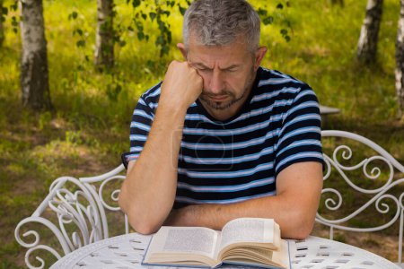 Téléchargez les photos : Un homme lit un livre dehors à une table. Il se repose. Concentration sélective douce. - en image libre de droit