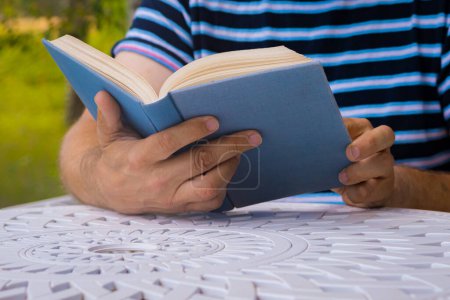 Téléchargez les photos : Un homme lit un livre dehors à une table. Il se repose. Concentration sélective douce. - en image libre de droit