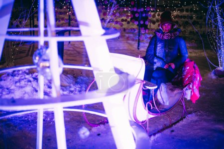 Foto de Mujer en trineo de renos y luces navideñas con árboles nevados, relajándose en vacaciones. Enfoque selectivo suave. Grano creado artificialmente para la imagen - Imagen libre de derechos