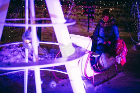 Foto de Mujer en trineo de renos y luces navideñas con árboles nevados, relajándose en vacaciones. Enfoque selectivo suave. Grano creado artificialmente para la imagen - Imagen libre de derechos