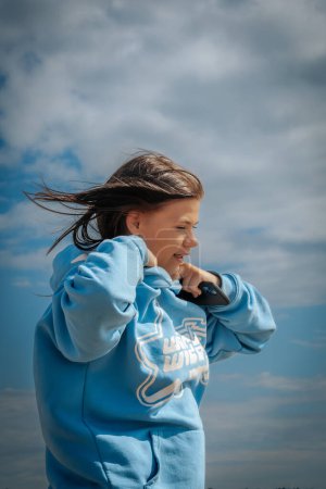 A young girl with brown hair wears a blue hoodie and holds a phone while enjoying a cool day outside under a partly cloudy sky.