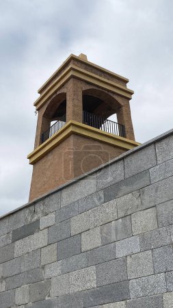 Una pared de hormigón texturizado se destaca sobre un telón de fondo de una torre de ladrillo marrón.