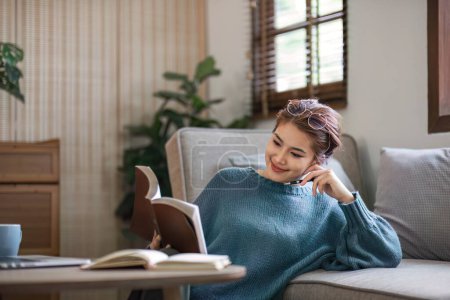 Téléchargez les photos : Jeune femme lecture livre sur confortable canapé gris contre à la maison.. - en image libre de droit