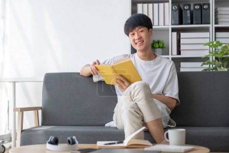 Photo for A happy young Asian man in casual clothes is sipping his morning coffee and reading a book on a sofa in the living room. Lifestyle concept.. - Royalty Free Image