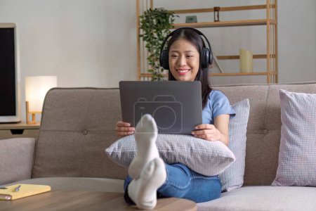 Foto de Atractiva joven feliz mujer escuchando música en los auriculares con placer en la sala de estar, tumbado en el sofá, utilizando el ordenador portátil y el descanso... - Imagen libre de derechos
