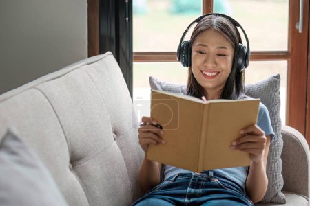 Foto de Mujer asiática joven escuchando música de auriculares y escribiendo nota para su idea de trabajo en el diario book.She sentado en el sofá gris en la sala de estar... - Imagen libre de derechos